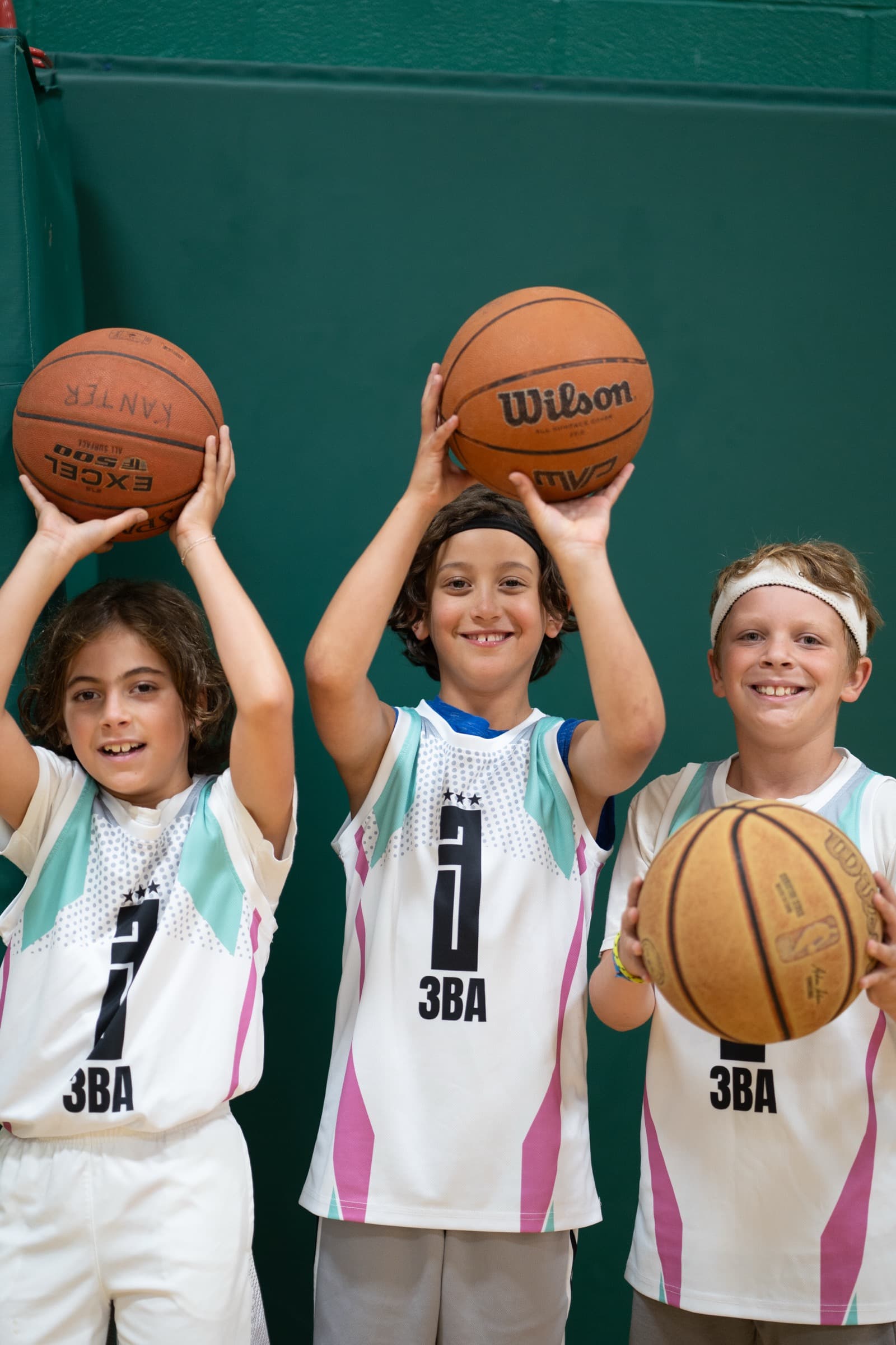 Three young players in white 3BA jerseys holding basketballs above their heads