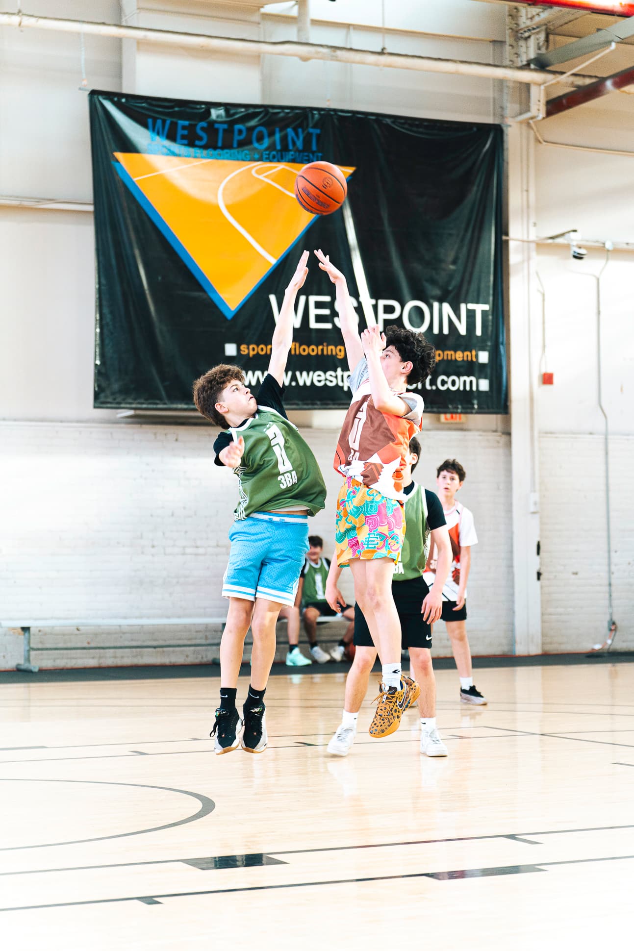 A player rising for a jump shot with a defender contesting, under the gym banners