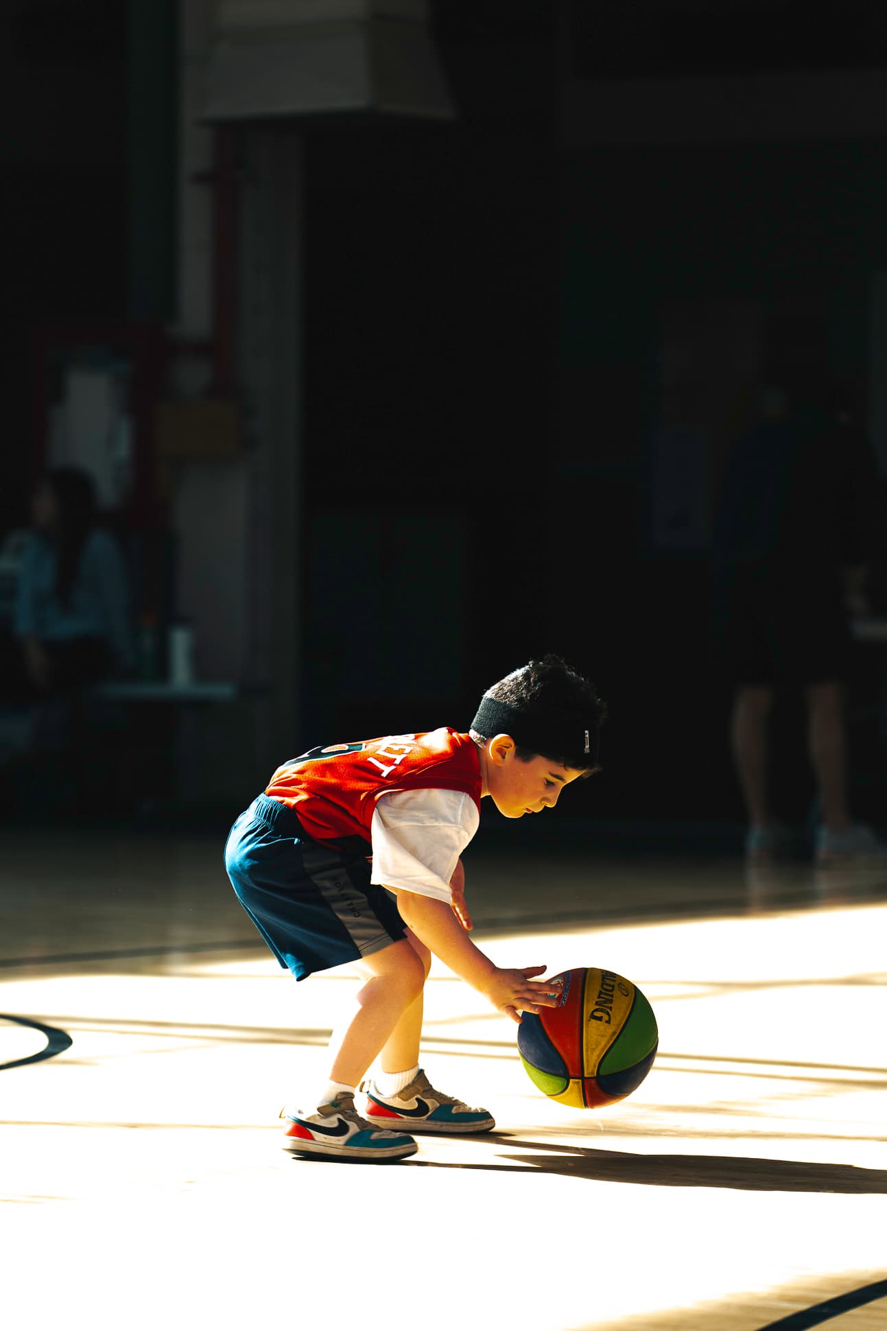 A young child bending down to pick up a basketball in a shaft of gym light