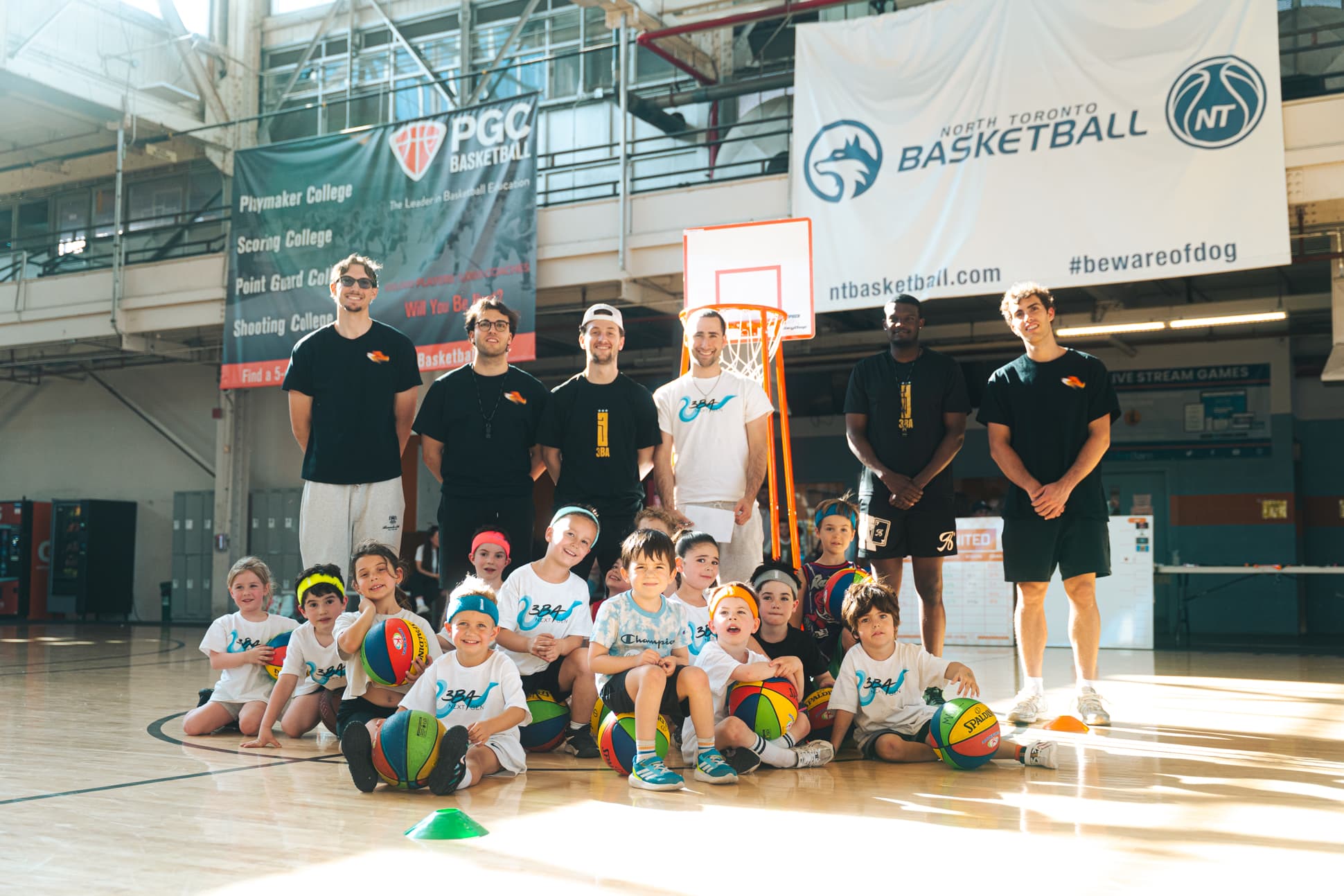 The 3BA coaching staff standing behind a group of young athletes seated on the gym floor with basketballs